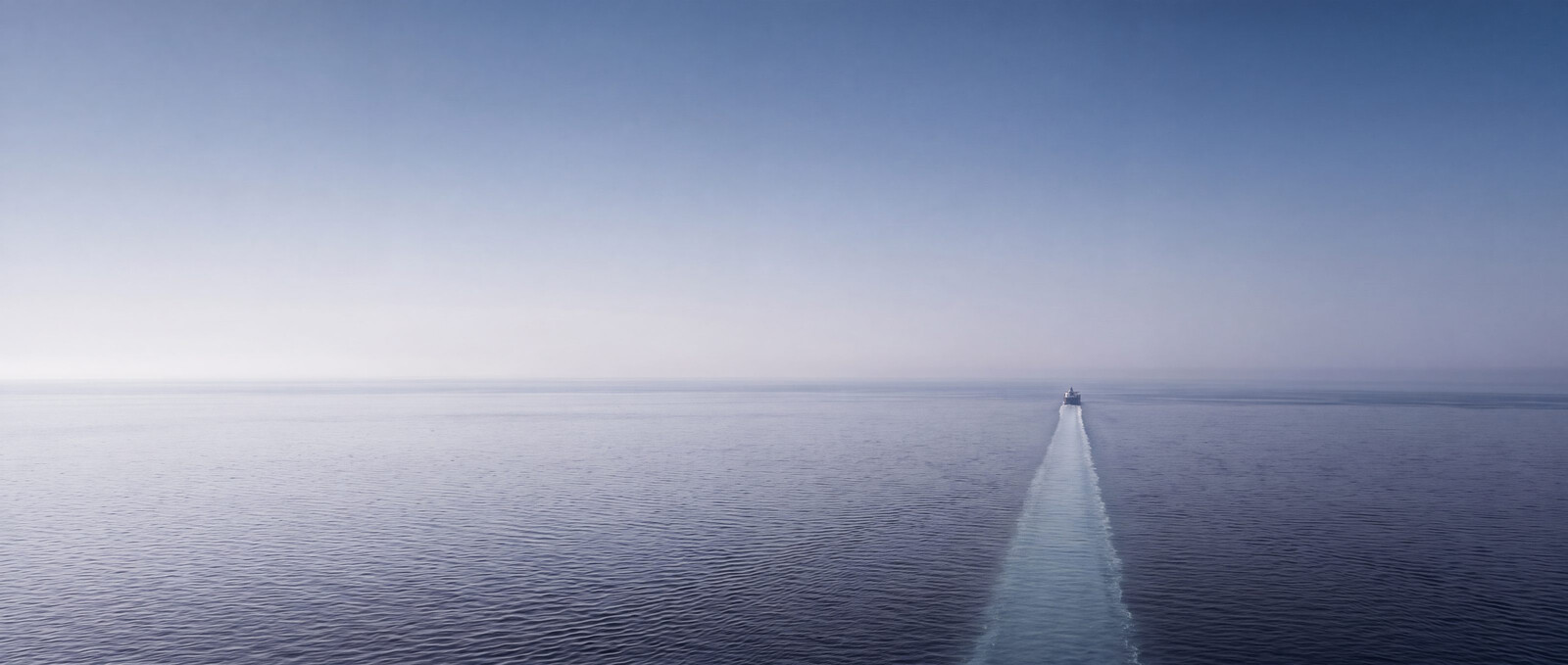 A lone bulk carrier holds a dead straight course toward the horizon at blue hour, her wake one straight line across a calm sea